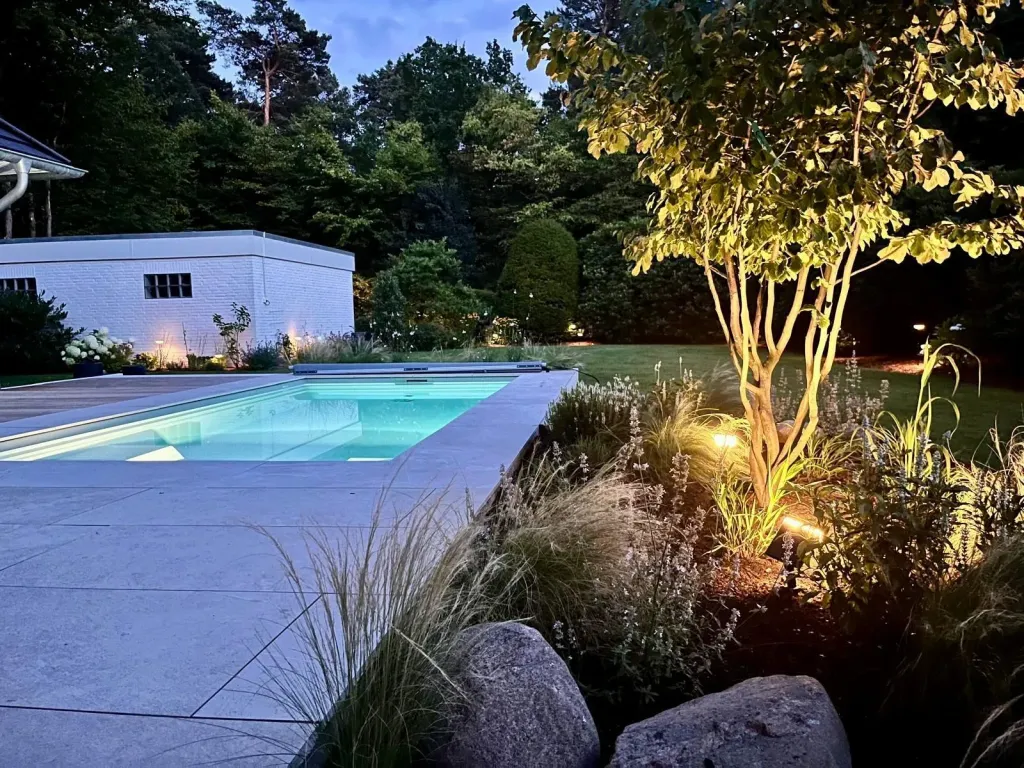 Beleuchteter Pool am Abend mit Ziergräsern und Findlingen